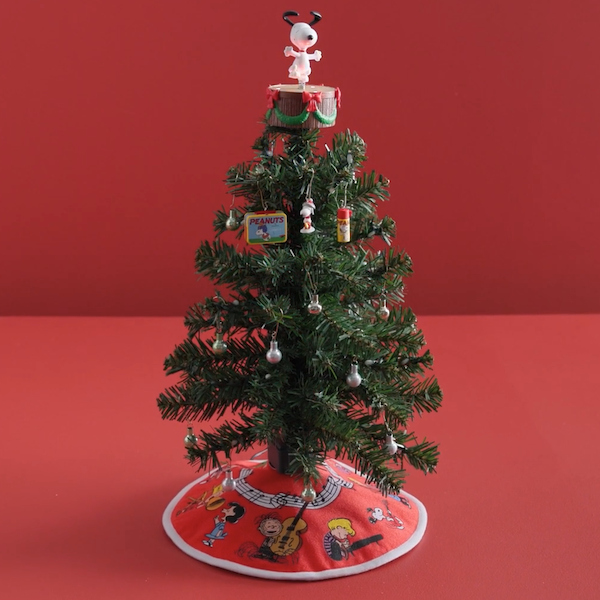 A miniature Christmas tree covered in mini Peanuts®-themed ornaments sits on a red surface against a coordinating red background; the tree has a mini tree skirt around its base that features all the characters of the Peanuts Gang, as well as a mini tree topper featuring Snoopy, dancing on a stage.